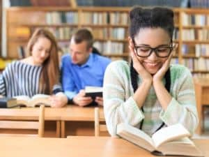 Smiling student wearing glasses reading a book in a library, with two other students studying together in the background.