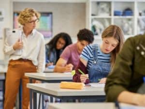 Teacher monitoring students as they complete a classroom writing assignment, with a girl in the foreground focused on her work while classmates study in the background.