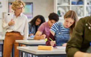 Teacher monitoring students as they complete a classroom writing assignment, with a girl in the foreground focused on her work while classmates study in the background.