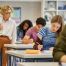 Teacher monitoring students as they complete a classroom writing assignment, with a girl in the foreground focused on her work while classmates study in the background.